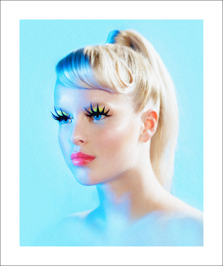Blond girl with blue eyes, fluor color pink lips, yellow eyeshadow and large black paper eyelashes is flooded with pastel blue light.