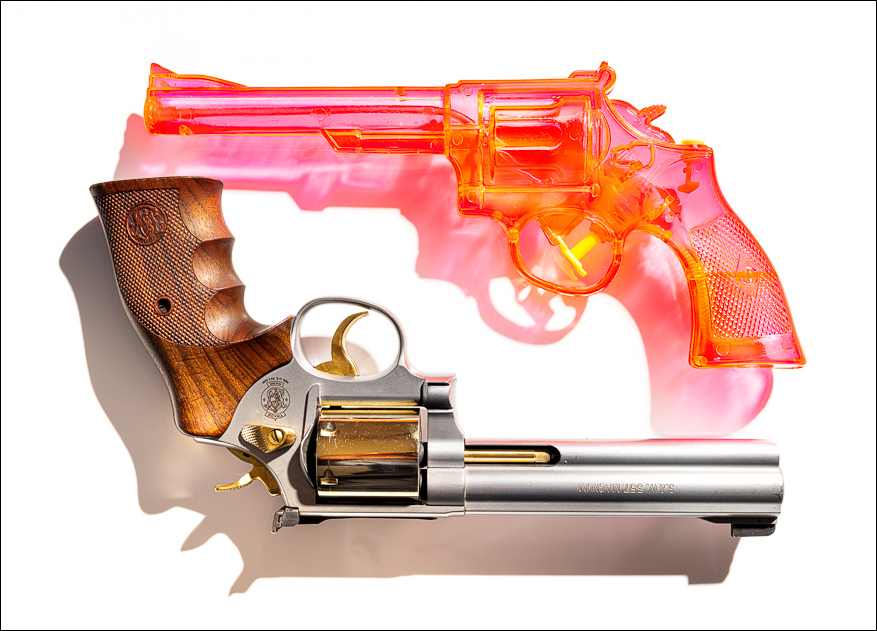 TOY AND TRIGGER: A pink translucent toy-like gun hovers above an upside-down Smith & Wesson Magnum revolver on a plain background.