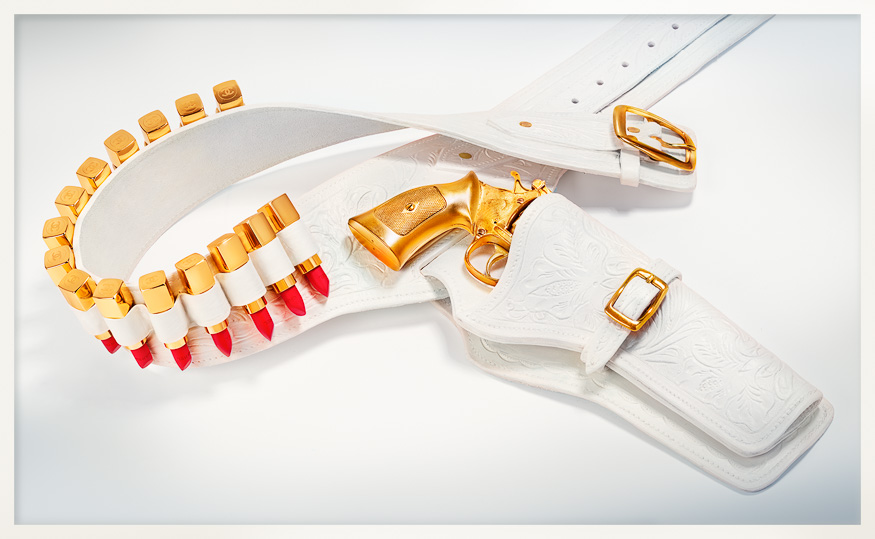 White leather holster belt with gold Chanel lipsticks and revolver against cool pink: an iconic fashionweapon of luxe and femme power.