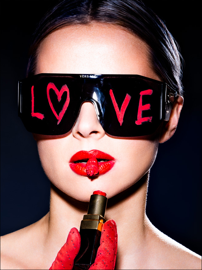 Bold girl with black sunglasses, 'LOVE' in lipstick, holds broken red lipstick and another piece between her lips, exuding confidence.