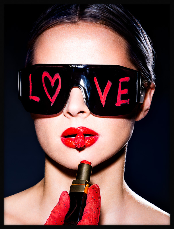 Bold girl with black sunglasses, 'LOVE' in lipstick, holds broken red lipstick and another piece between her lips, exuding confidence.